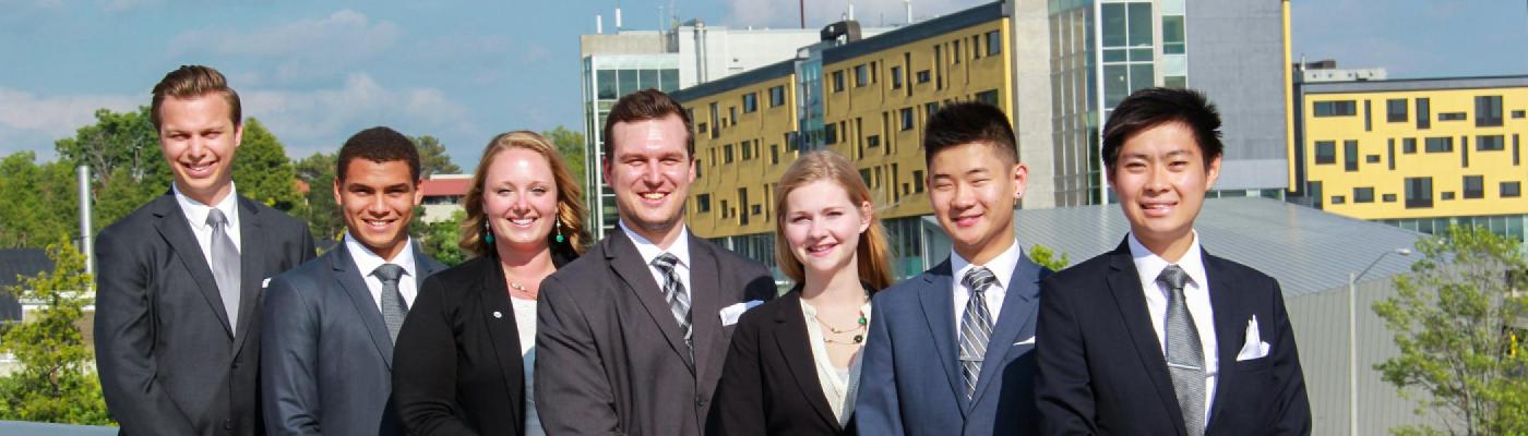 6 Students in business suits smiling at the camea standing on the Faryon bridge in the sunlight