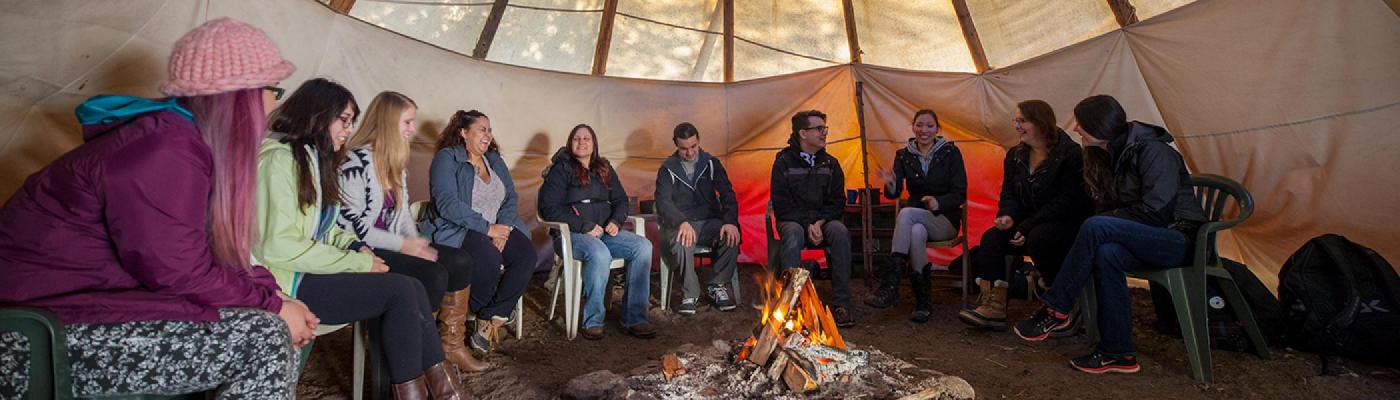 A group of students sitting in a circle in a teepee around a fire talking