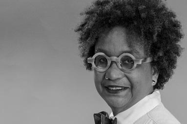 Black woman with curly hair, round glasses and dress shirt with bow tie facing camera and smiling