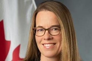 a smiling woman wearing glasses has straight blond, shoulder length hair. A Canadian flag on a mast is in the background.
