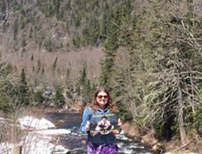 Person standing in front of river in wooded area, holding up poster for school for the Study of Canada