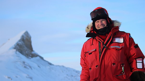 Man wearing red parka and snow gear smiling at camera from sunny Arctic landscape