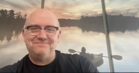 Mike Perry smiles at the camera in front of a painting depicting a kayak on a lake at sunrise.