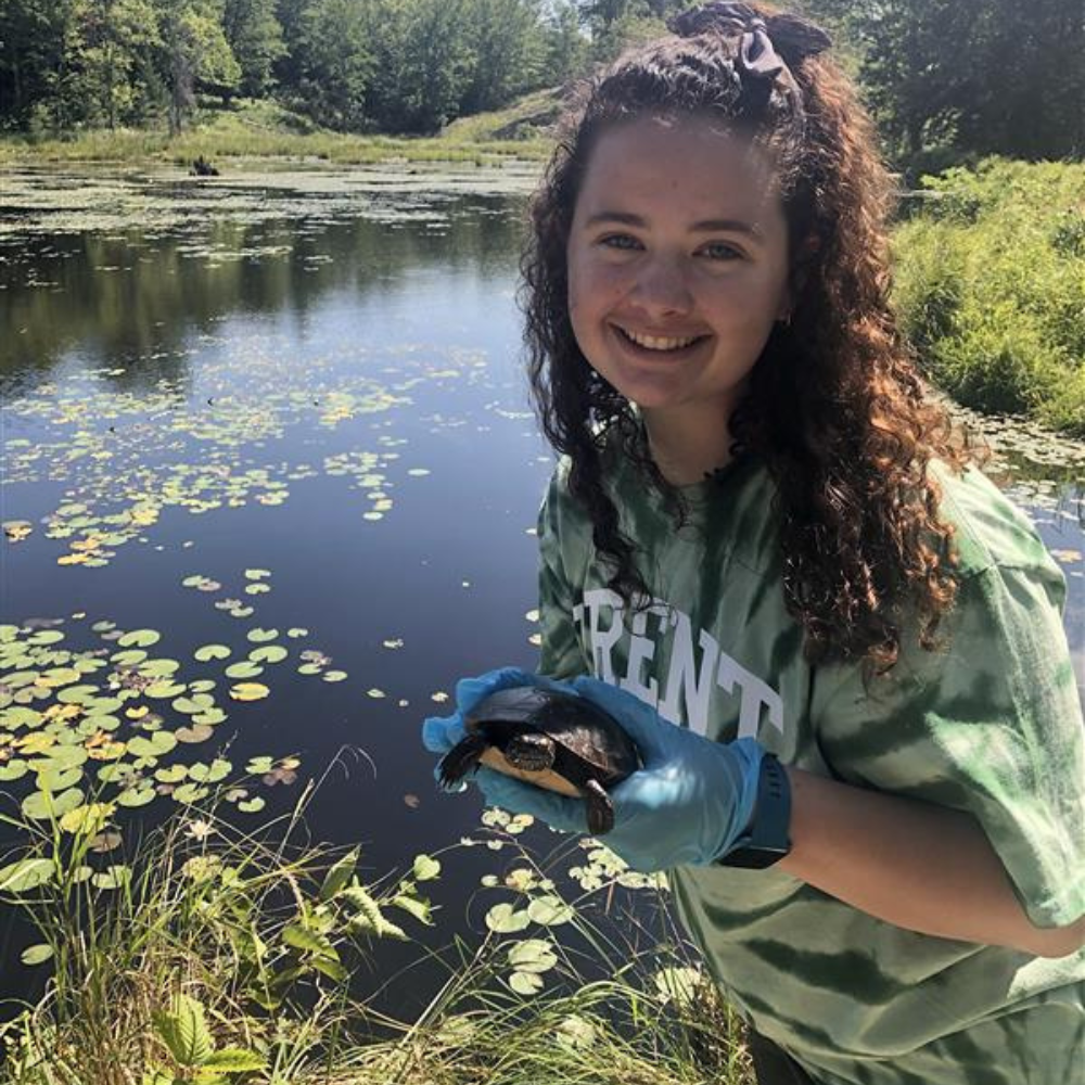 Student smiles, holding a turtle by a pond.