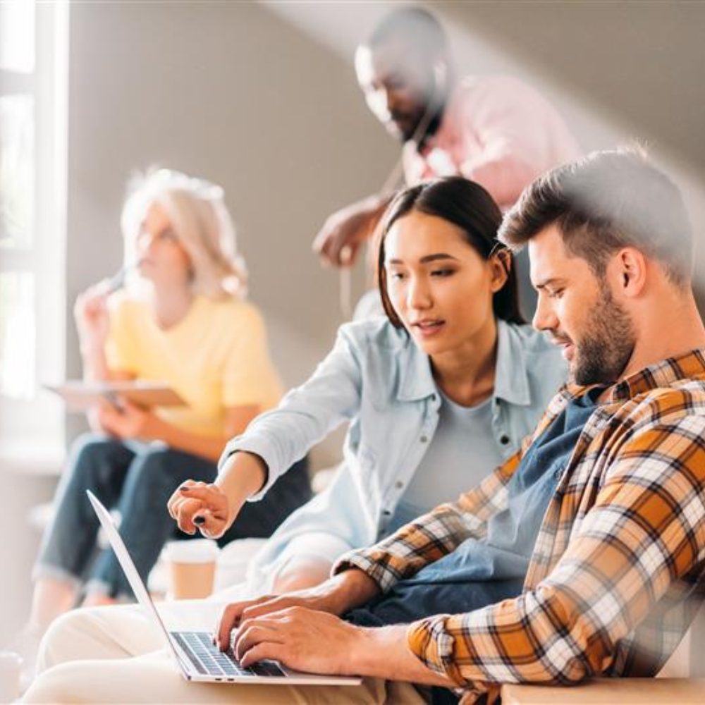 Four young professionals collaborating in a bright, modern workspace. A woman in a light blue shirt is pointing at a laptop screen while discussing something with a man in a plaid shirt who is typing.