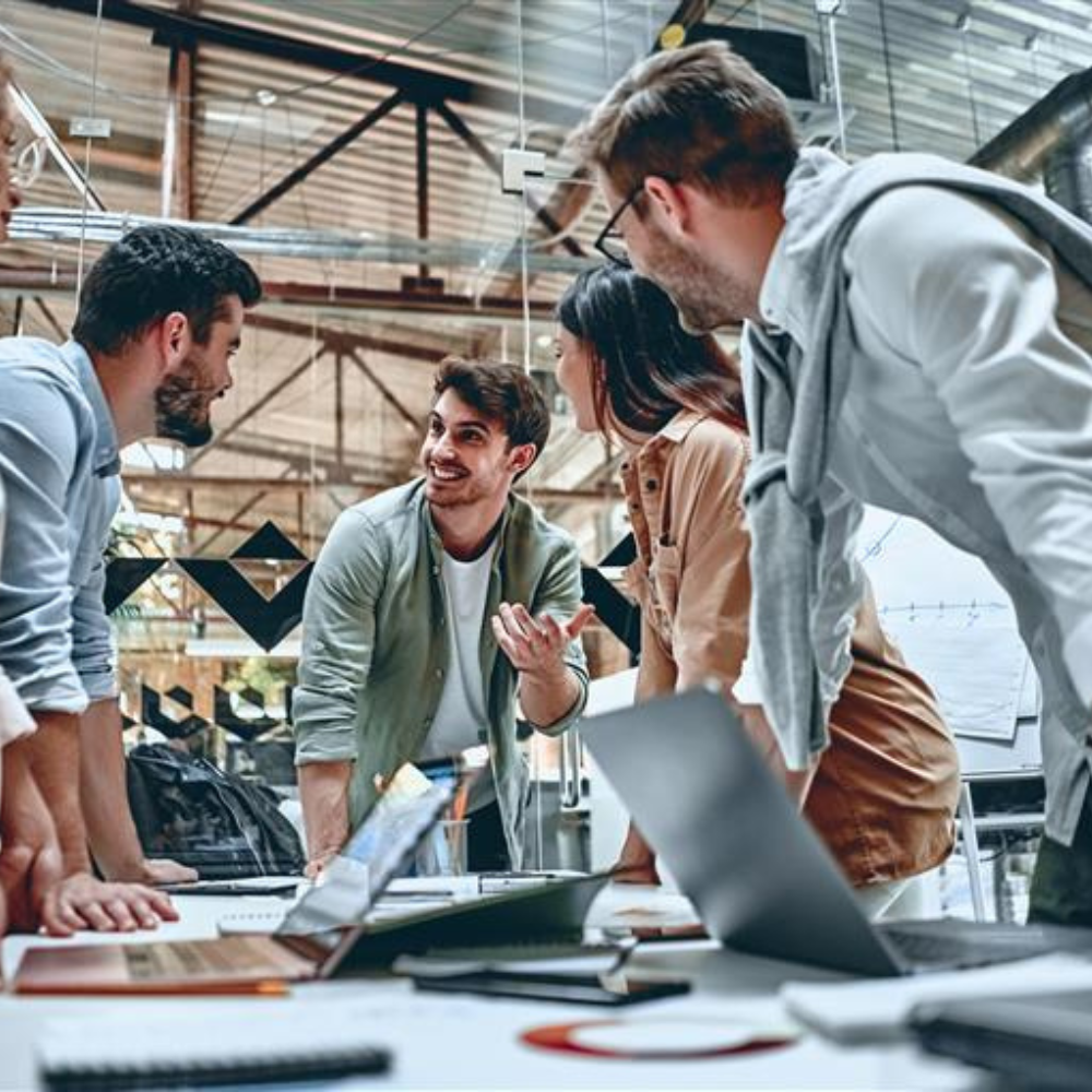 Group of five young professionals gathered around a table in a modern office, actively engaged in discussion. One man in the center is speaking and gesturing enthusiastically, while the others listen attentively.