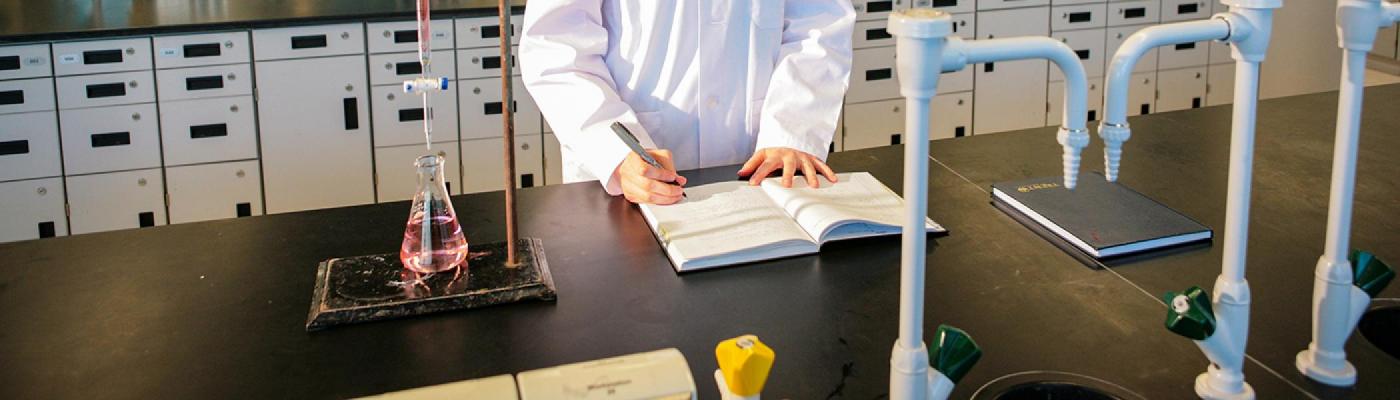 A chemistry student working at a chemistry lab bench, writing in a notebook beside a test tube with pink liquid in it