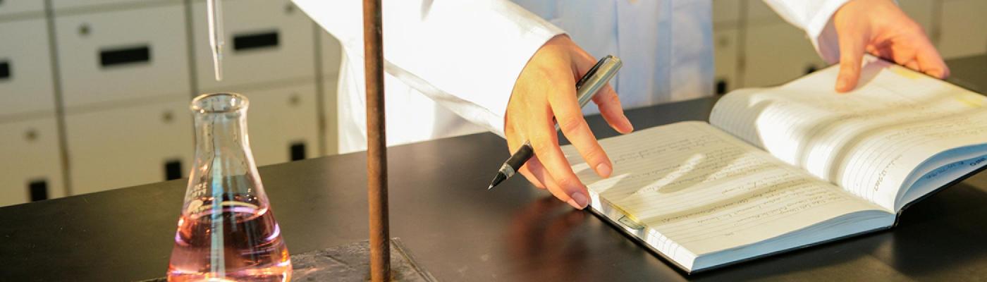 A pair of hands resting on a lab notebook, writing notes, in front of an Erlenmeyer flask of pink liquid and burette on a chemistry lab bench