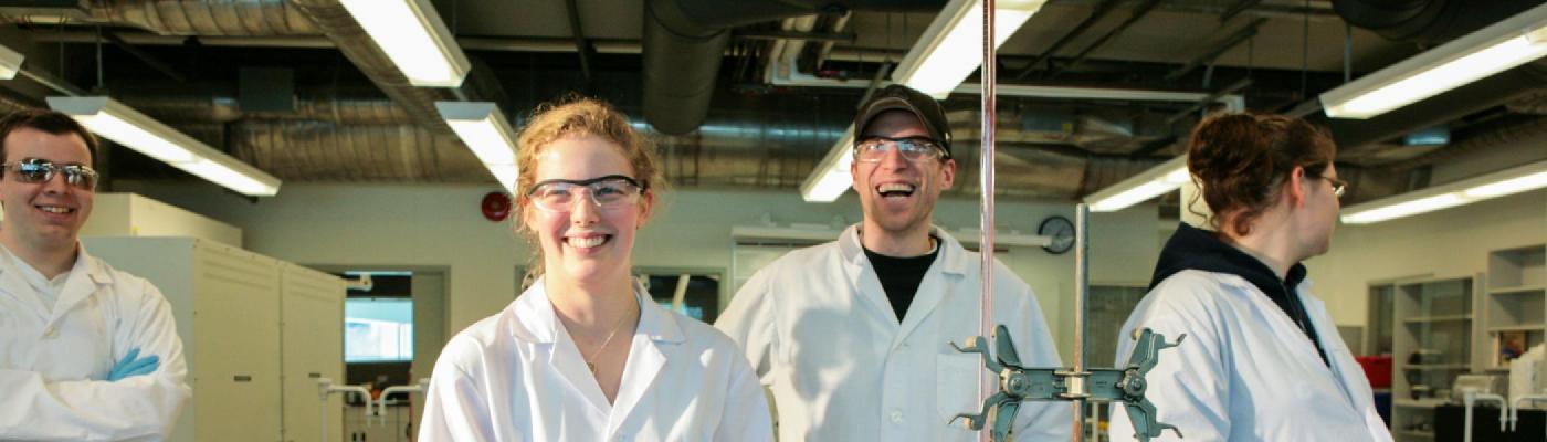 4 Chemistry students standing in a chemistry lab smiling at the camera, wearing white lab coats and safety goggles