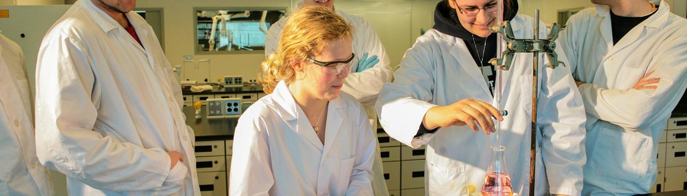 A group of 4 chemistry students standing around a chemistry workbench titrating chemicals using a burette and Erlenmeyer flask