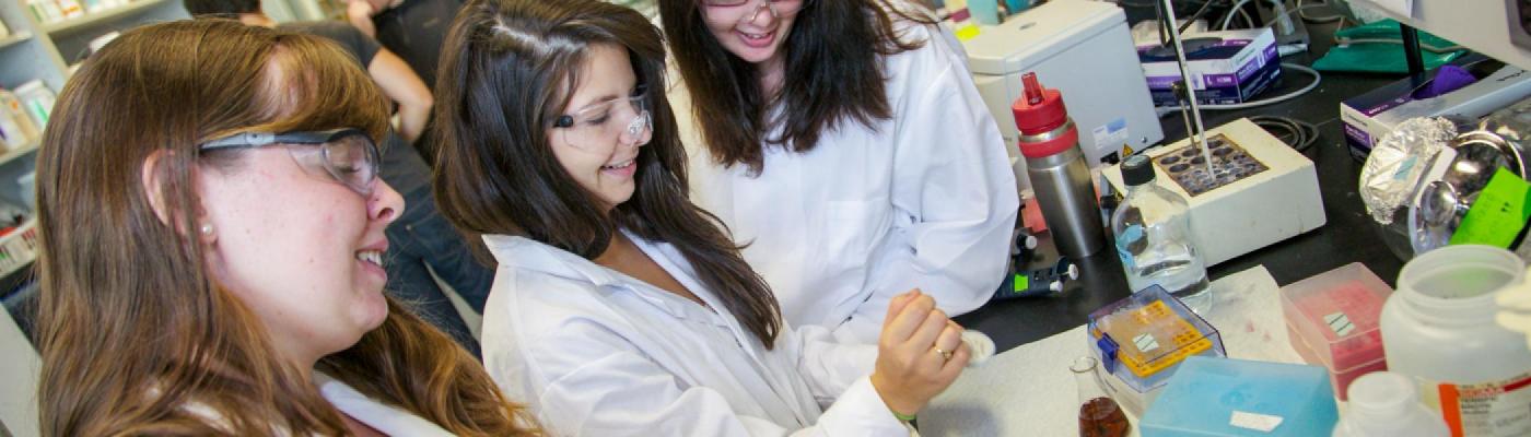 3 Biochemistry students standing at a lab desk mixing chemicals, in white lab coats and safety glasses