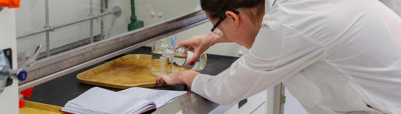 A female chemistry student working in the fumehood pouring a chemical from a bottle into a graduated cylinder, wearing a white lab coat and safety glasses