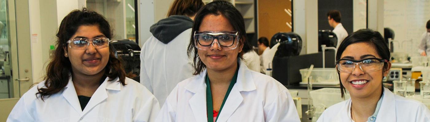 3 Chemistry students standing at a lab desk, in white lab coats, smiling at the camera