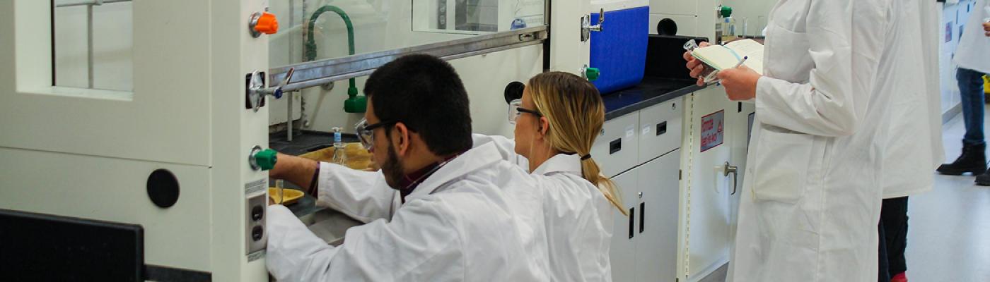 2 Chemistry students kneeling at a chemistry lab fumehood getting chemicals, wearing white labcoats and safety goggles, a 3rd student waits behind
