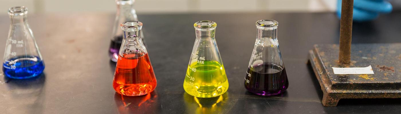 Five chemistry Erlenmeyer flasks on a black chemistry lab bench with different coloured liquids in them