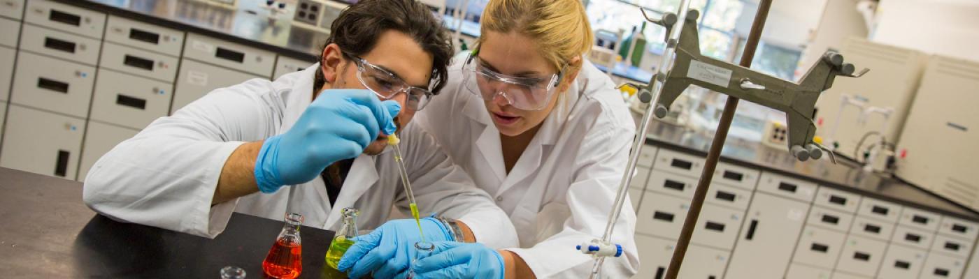 2 Chemistry students working with coloured solutions in Erlenmeyer flasks and glass pipettes in a chemistry lab, wearing white lab coats and safety glasses