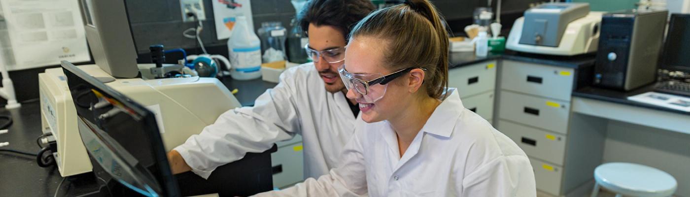 2 Chemistry students looking at a computer screen in a chemistry lab beside an FTIR instrument wearing white lab coats and safety glasses