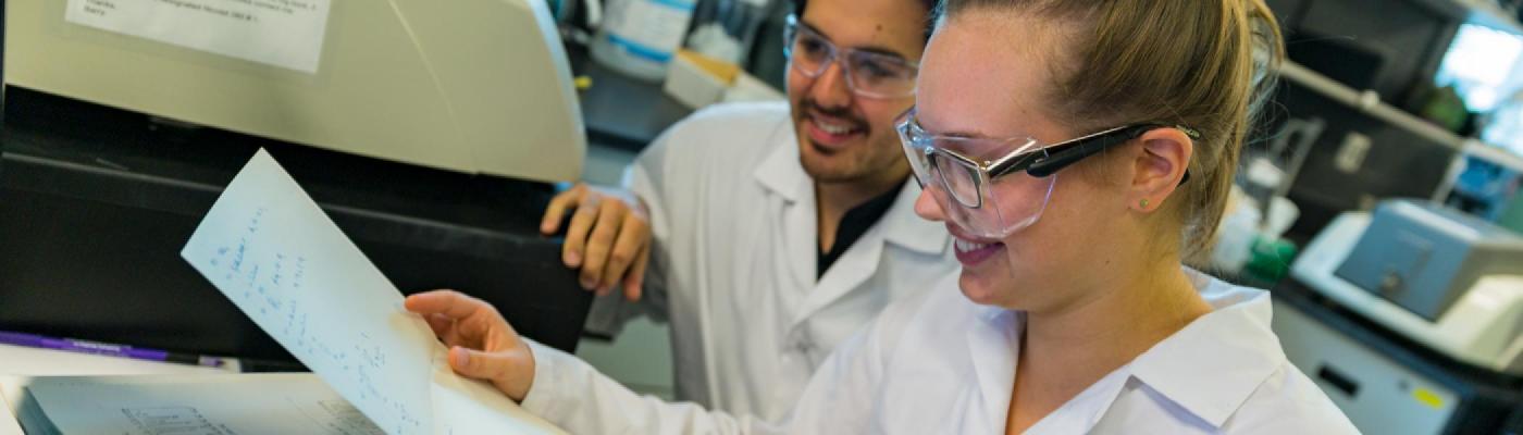 2 Chemistry students sitting at a computer in the instrumental chemistry lab looking at a paper, wearing white lab coats and safety goggles