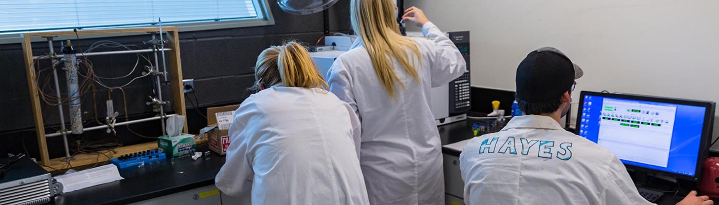 3 Chemistry students working in a chemistry lab on a GC-MS instrument and computer, wearing white lab coats