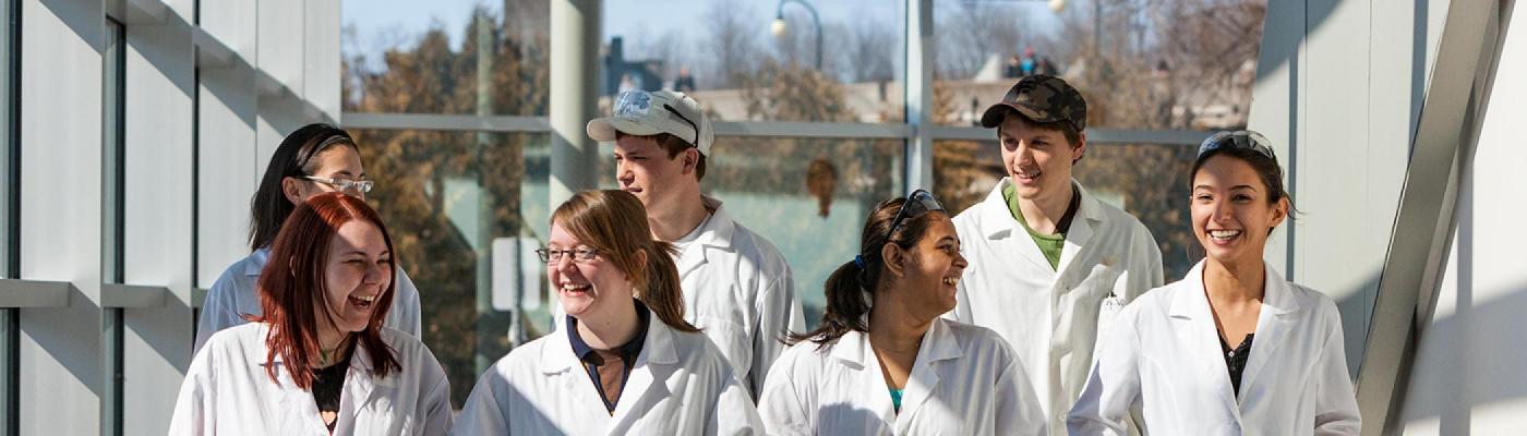 A group of 7 chemistry students walking in a hall smiling and talking, wearing white lab coats
