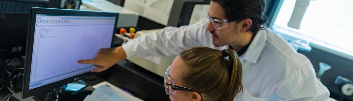 2 Chemistry students looking at an IR spectrum on a computer screen in a chemistry lab wearing white lab coats and safety glasses