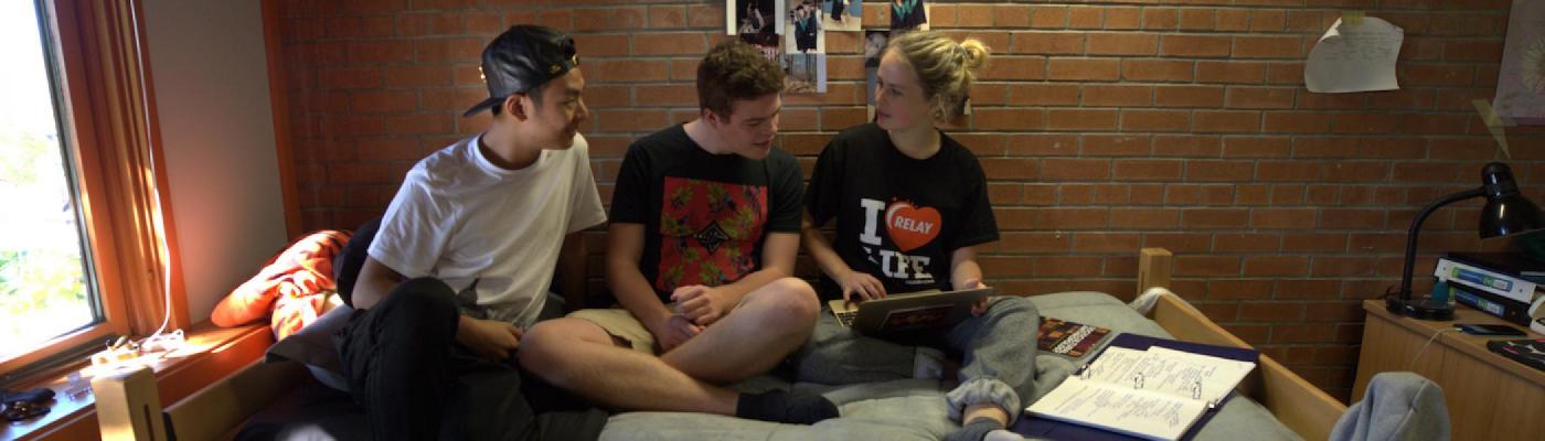 2 young men and a young woman sitting on a bed together in a residence room looking at a laptop