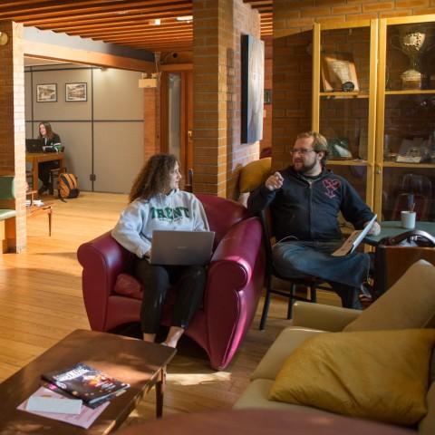 Two students have a conversation in Traill College's dining space, The Trend. The space is decorated in a pub-style with warm lighting, a mix of plush seats and dining tables, a coffee table with reading materials, and a display case of memorabilia.  