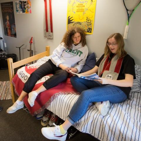 Two students studying on a bed in Traill College's residence. There are several decorations on the wall behind them including a tennis racquet, posters, and a Traill College scarf.
