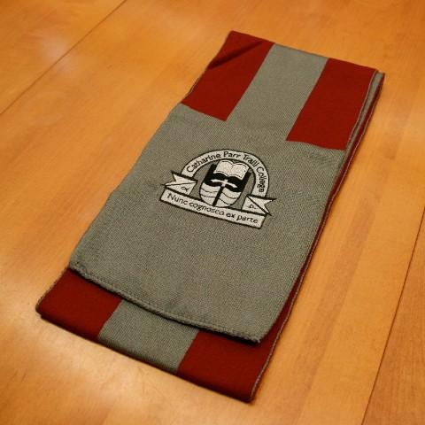 A grey Traill College scarf with a wide maroon stripe running along each side. The Traill College logo is embroidered on the end of the scarf, which is sold grey.