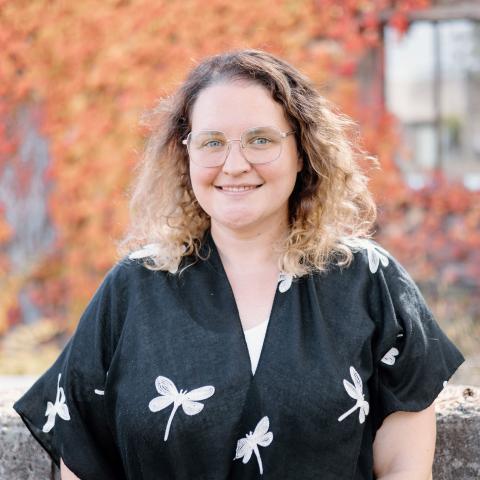 Jackie Orsetto. She is smiling and standing in front of a tree with orange autumn leaves. She is wearing a black shirt with white dragonflies on it.