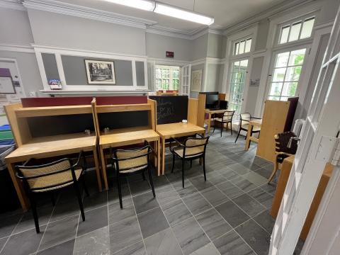 The Greg Piasetzki Reading Room in Scott House. The walls and tile floors are grey and several study cubicles made of warm-toned wood and maroon accents are arranged around the room. There are large windows along the right side of the room and a fireplace mantle with artwork above it towards the back of the room.