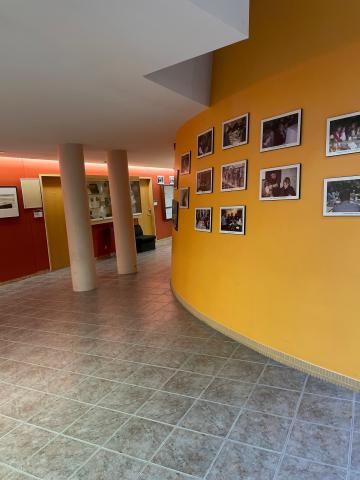 A photo of the second floor of Otonabee College - where a golden wall is covered in images of students, staff and faculty from years past. The floor winds around a corner where there are two pillars.