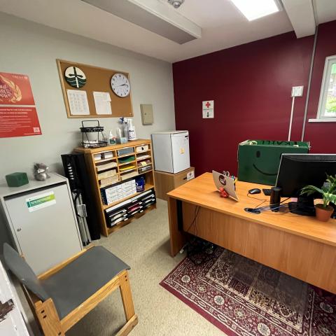 The Traill College Office in Scott House. A grey chair sits facing a wooden office desk. Sitting at the desk behind the computer is Traill's mascot, the Happy Box. It is a green cardboard box with a smiley face drawn on the front. Along the left side of the room are shelves of office supplies, a bulletin board, and a mini-fridge. 