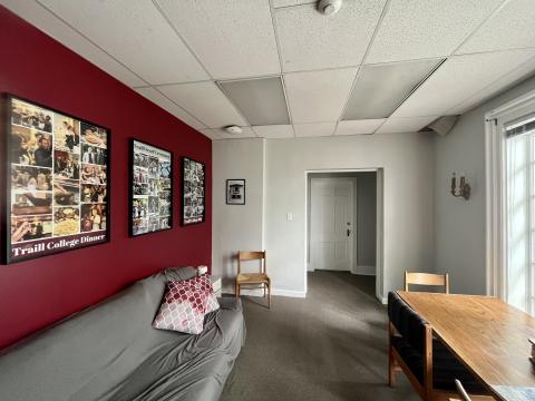 The second floor lounge in Scott House. The walls are maroon and grey, with photo collages of College events on the wall. There is a wooden table on the right side of the room and a grey couch with maroon cushions on the left.
