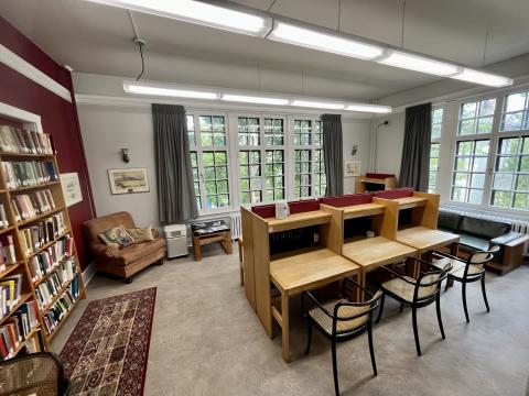 The Graduate Reading Room in Scott House. There are large windows along two walls, and along a third are large bookcases. There is a couch, a plush chair, and study cubicles arranged around the room.