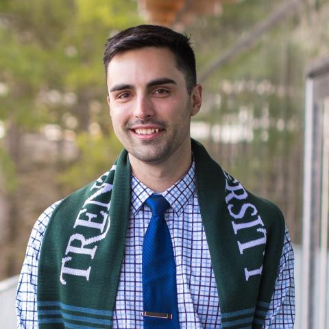 Zachary Brault. He is smiling and has a green Trent University scarf draped over his shoulders. He is wearing a blue checked dress shirt with a blue tie.