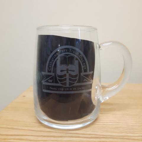 A clear glass mug. A black napkin has been placed inside the mug to show the opaque Traill College logo etched into the glass.