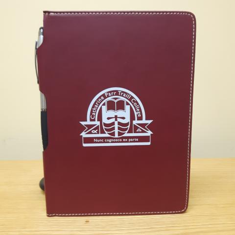 A rectangular notebook with a maroon, faux-leather cover. There is a pen clipped to the side of the notebook and a white Traill College logo in the centre.