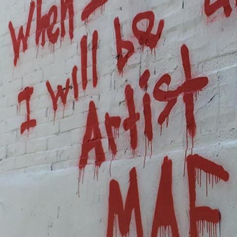 Words painted with red spray paint onto a white brick wall. A portion of the graffiti is cut off, but the words that are in the photo are, 
"when", "I will be", and "artist".
