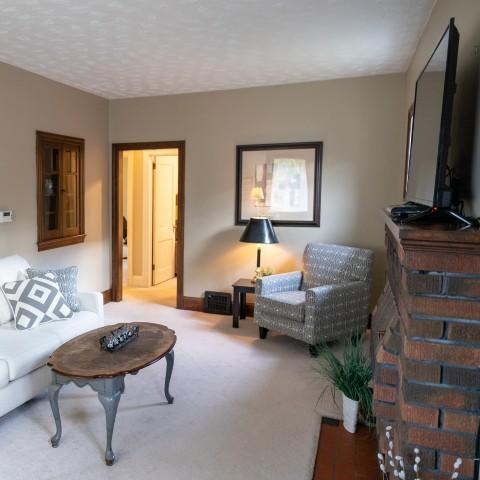 The living room of a guest suite at Traill College. The walls and carpet are off-white. There are a sofa, chair, coffee table, television, and brick fireplace.