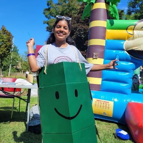 A Traill student wearing Traill College's Happy Box mascot costume. The costume is made from a cardboard box and painted green with a smiley face drawn in the centre. The box is hanging from the student's shoulders with twine rope. The student is at the Traill College Carnival, standing in front of a colourful bouncy castle.