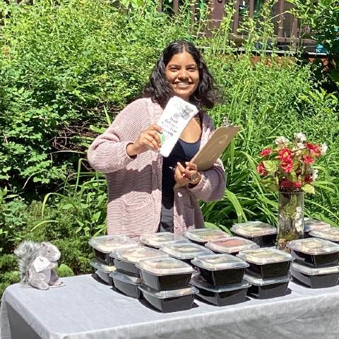 A student standing behind a table at the Traill College Teddy Bear Picnic. The table is full of catered lunch containers and the student is holding an event brochure.