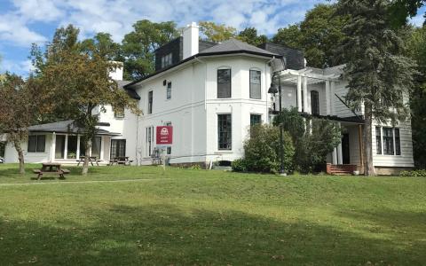 The green lawn outside Scott House at Traill College. Scott House is a large, white Victorian-style home with a dark grey roof. There are bushes, two fruit trees, and a picnic table outside the house.