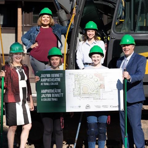 Marilyn Burns (left), Michael Eamon (right), and other Traill staff at the ground breaking ceremony for the Jalynn Bennett Amphitheatre. They are wearing green hard hats and posing in front of construction equipment.