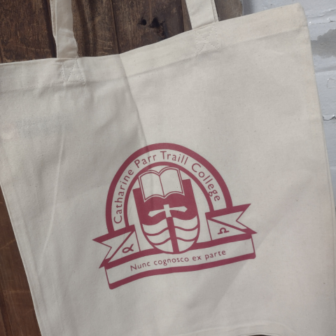 An off-white cotton tote bag with a maroon Traill College logo