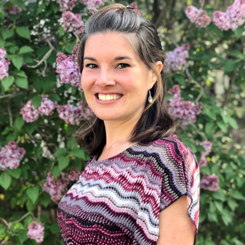Samantha Perritt. She is standing in front of a lilac bush and smiling. She has her hair in a half-up-half-down style and is wearing a striped purple shirt.