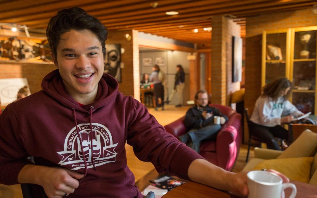 A student enjoys a coffee in Traill College's dining space, The Trend. The student is wearing a maroon Traill College Hoodie.