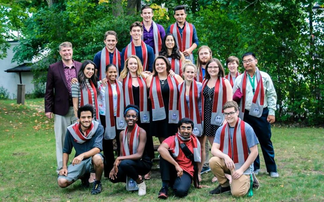 A group of Traill students and the Traill College Principal wearing matching Traill College scarves. They are smiling and posing in front of a background of green grass and a large tree.