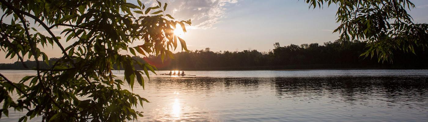 The sun coimng up on the Otonabee river, between trees on the river bank while a team of rowers rows a boat along the river