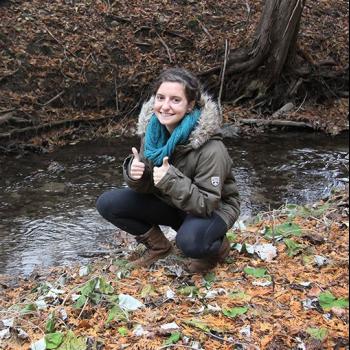 Student researcher crouching next to creek and smiling with thumbs up.